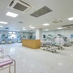Spacious hospital NICU with multiple infant incubators, monitors, and pink bassinets along the walls; a wooden reception desk in the center.