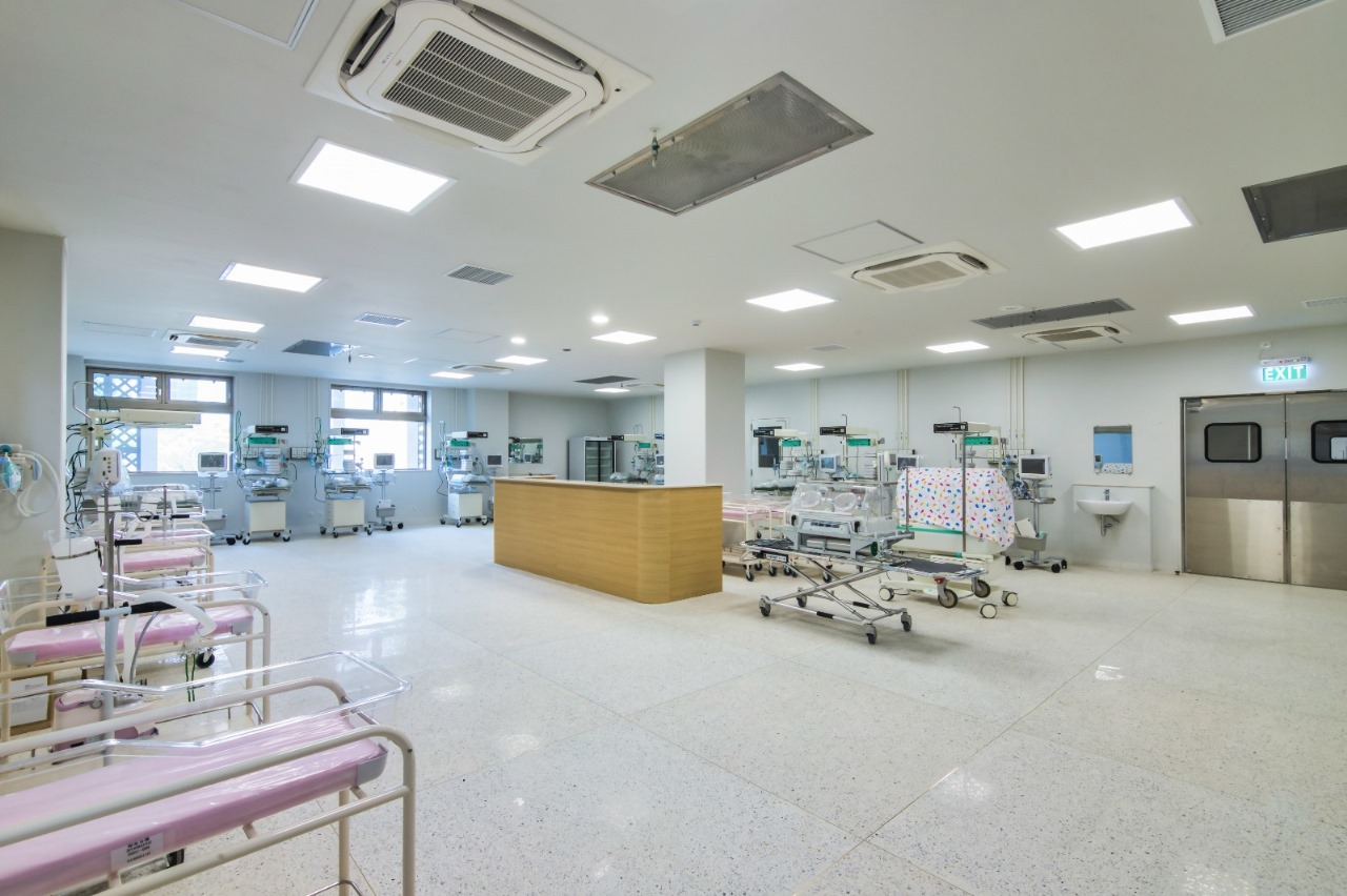Spacious hospital NICU with multiple infant incubators, monitors, and pink bassinets along the walls; a wooden reception desk in the center.